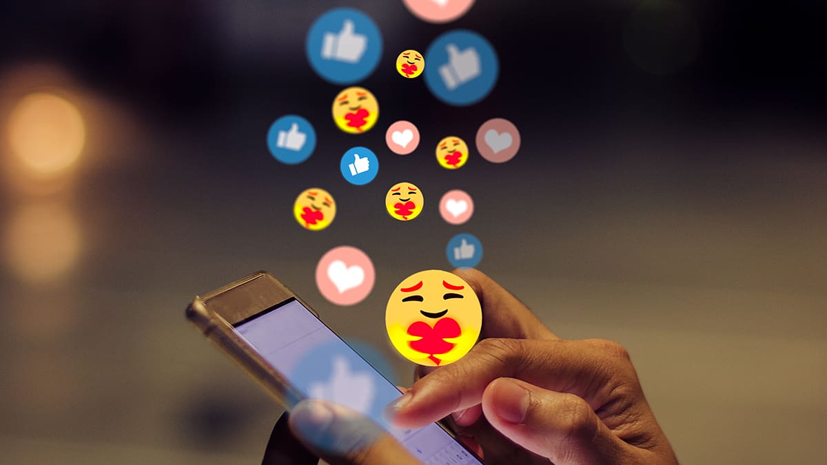 Image of a mobile phone user interacting with social media by reacting to a post. Like, love, and care emojis are shown above phone to illustrate this engagement.