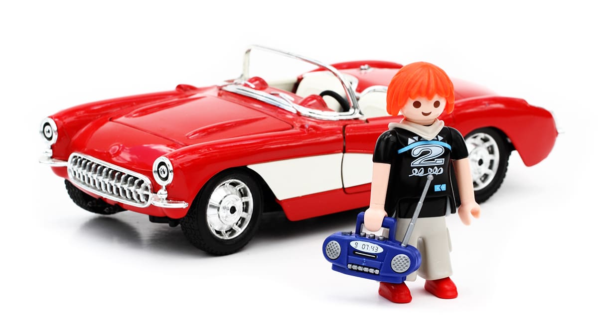 A young adult toy figure carrying a boombox and standing in front of a toy sports car.